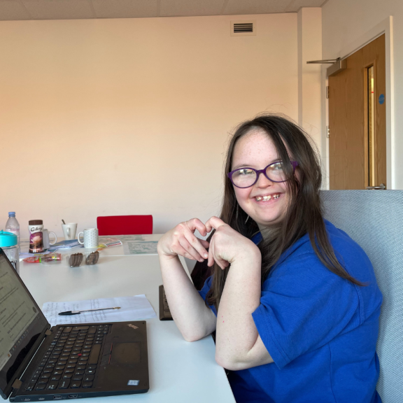 youth council member sitting at a desk with a laptop making a heart symbol with her hands 