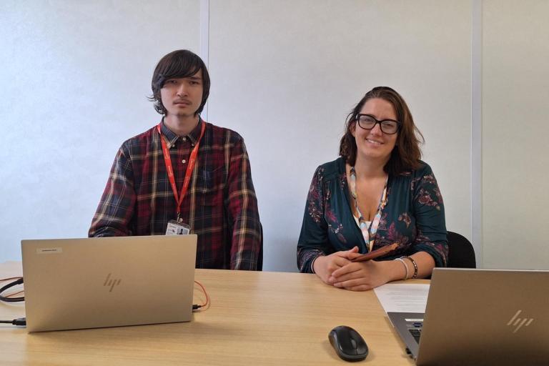 two people smiling at a desk with laptops 